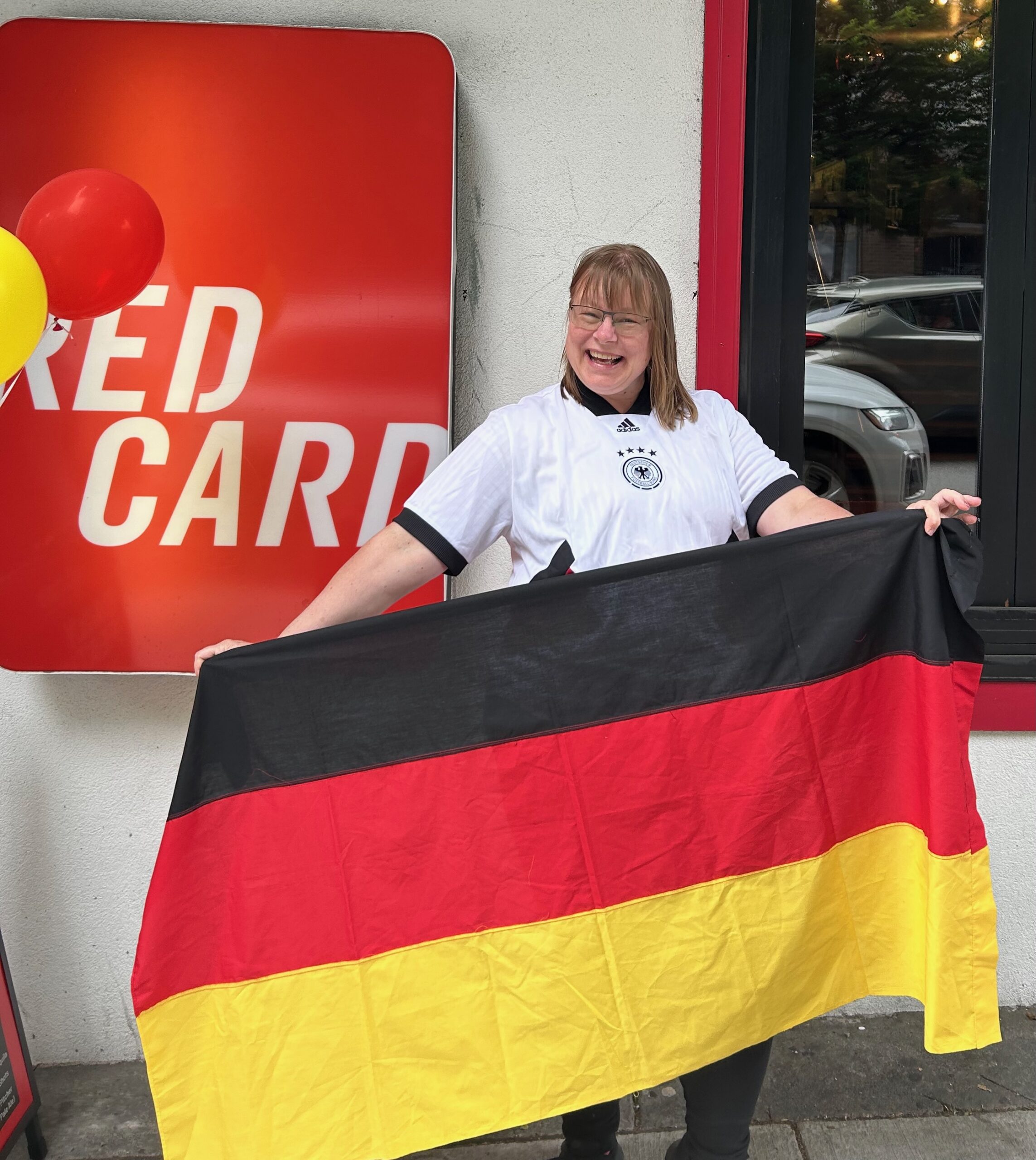 Elke holding the German flag in front of the Red Card Bar & Grill in 2024